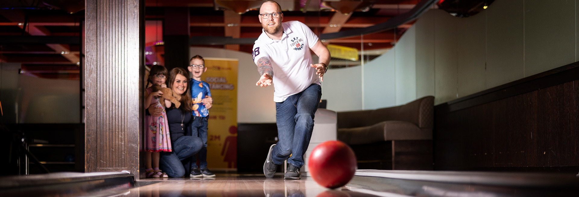 Father rolling a red bowling ball down a lane whilst wife and children watch and smile